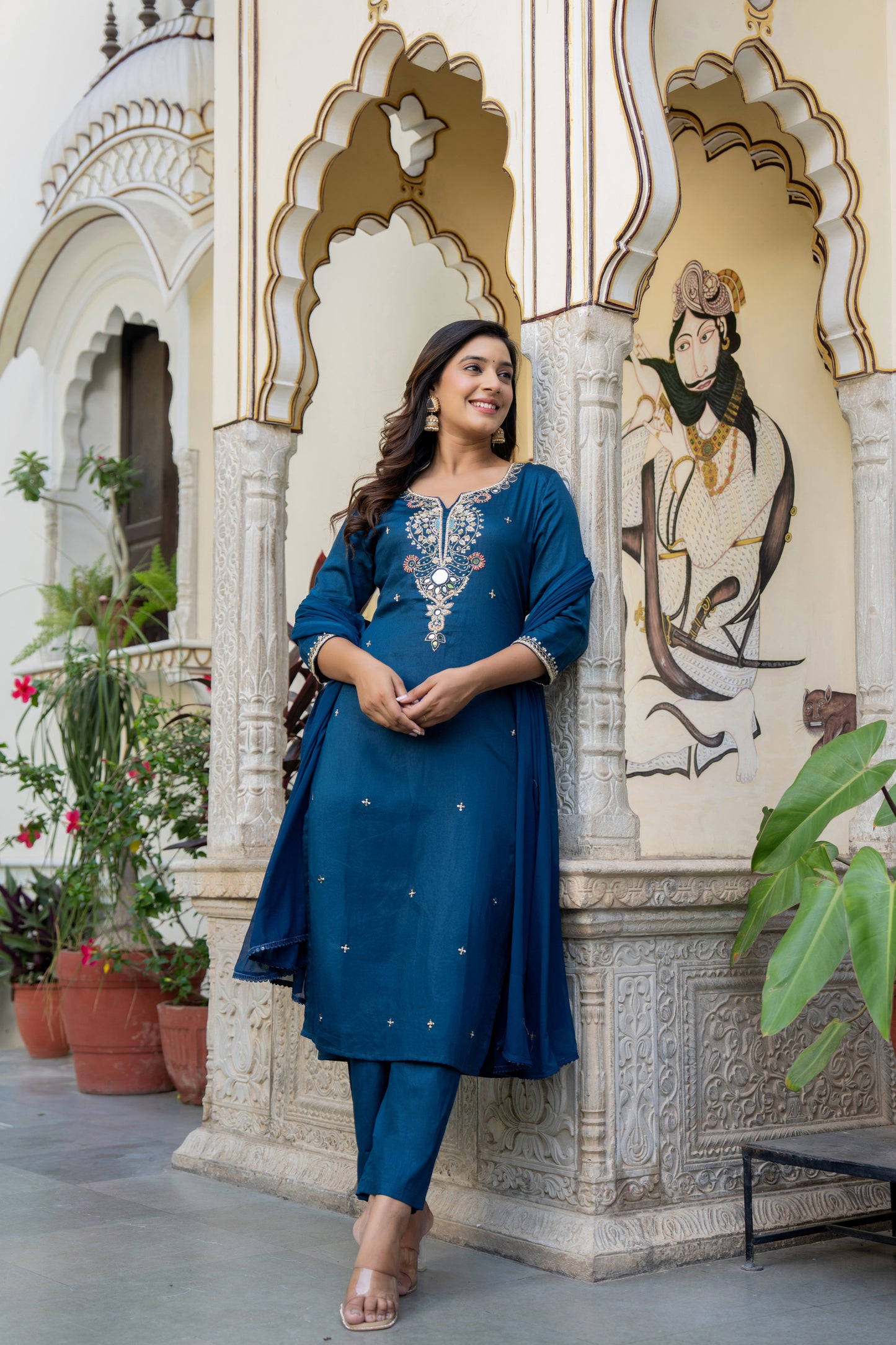 Woman in a Teal blue Kurta set made for wedding wear standing in front of ornate architectural wall.