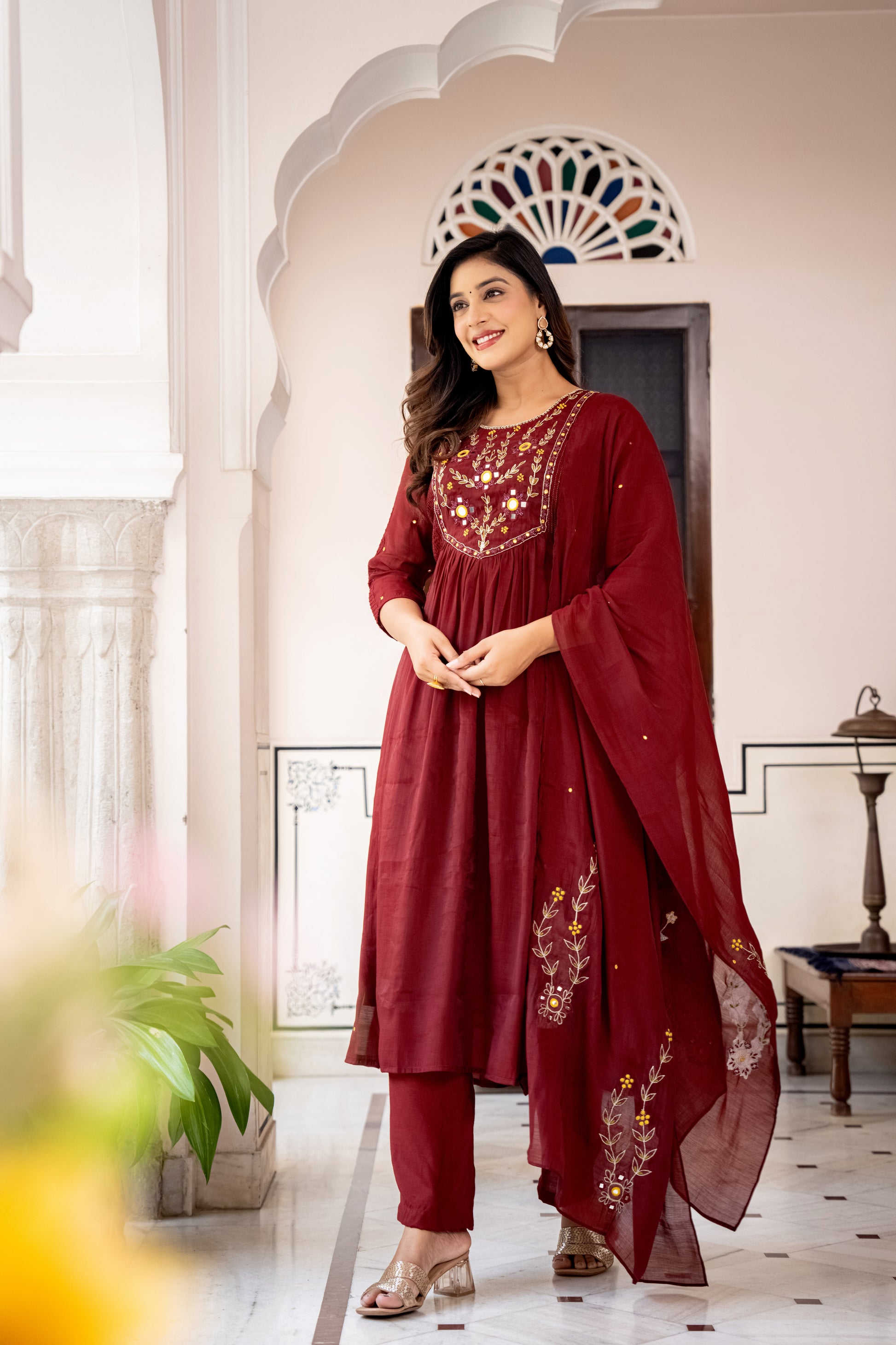 Woman in a red Kurta set with embroidery designed for wedding standing in a decorated room.