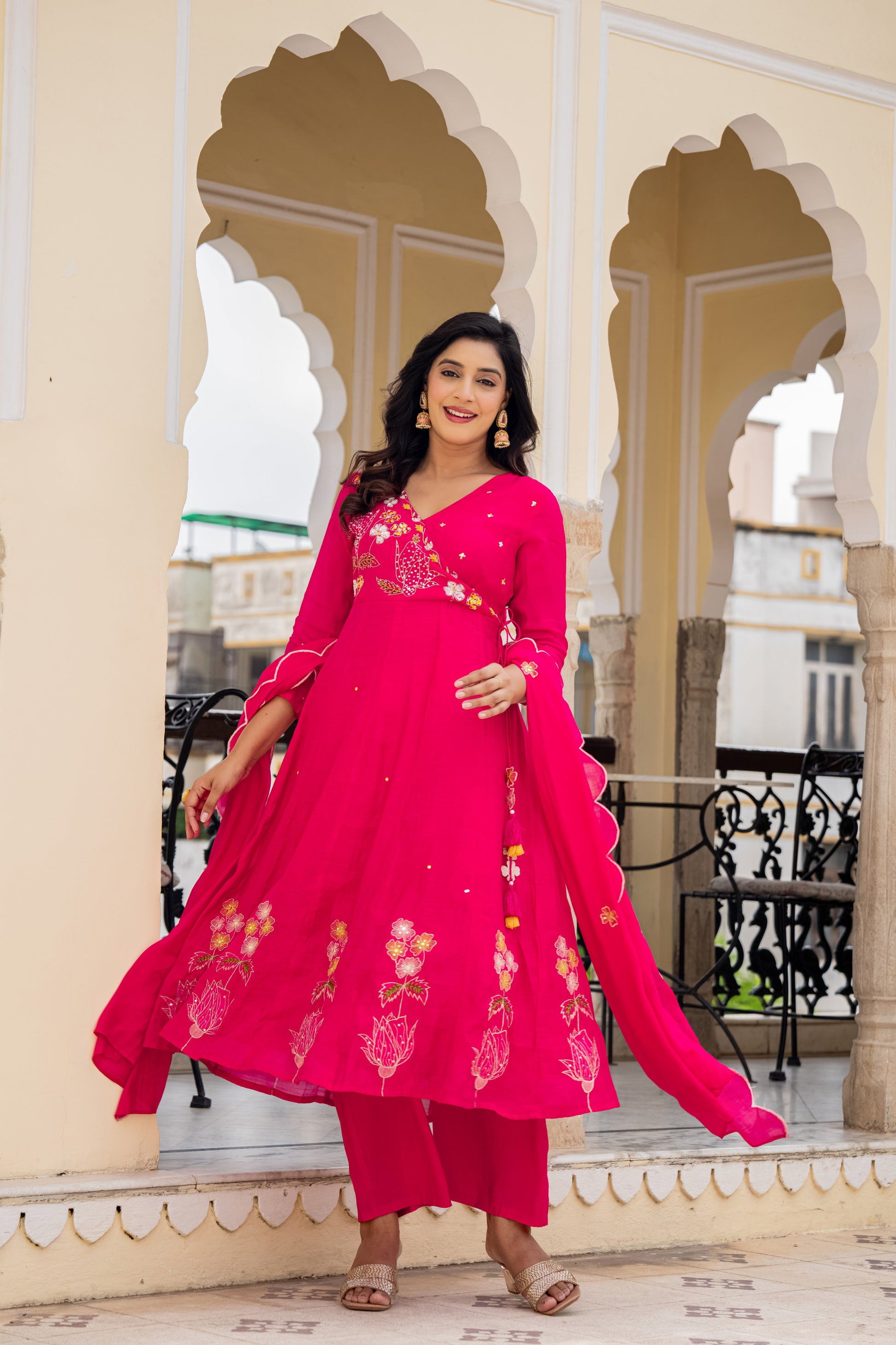 Woman in a bright pink anarkali kurta set with dupatta standing in front of ornate arches, showcasing festive kurta sets for women