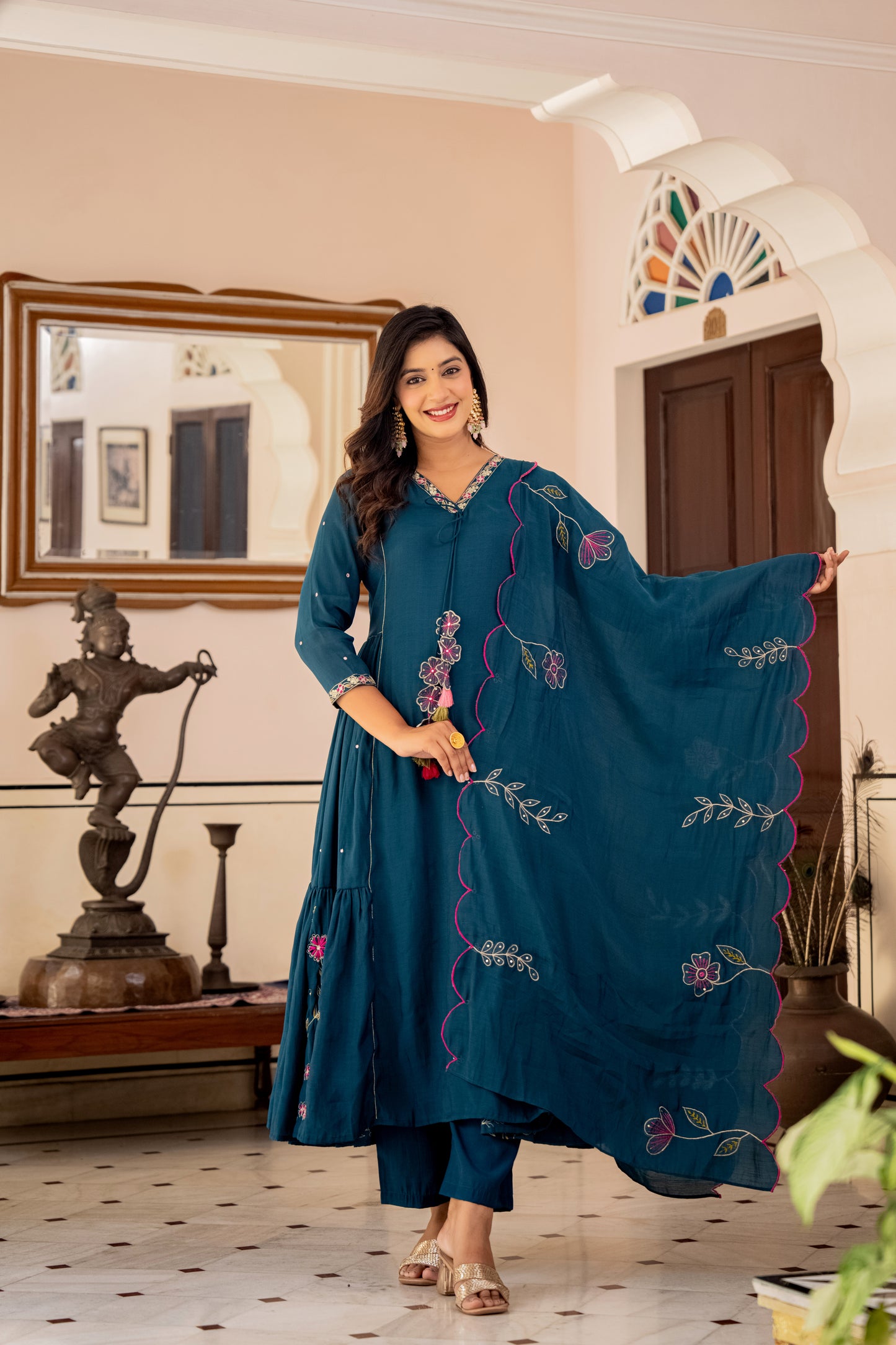 Woman in a teal embroidered outfit standing in a room with decor elements.