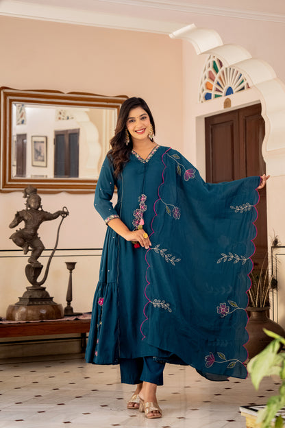 Woman in a teal embroidered outfit standing in a room with decor elements.