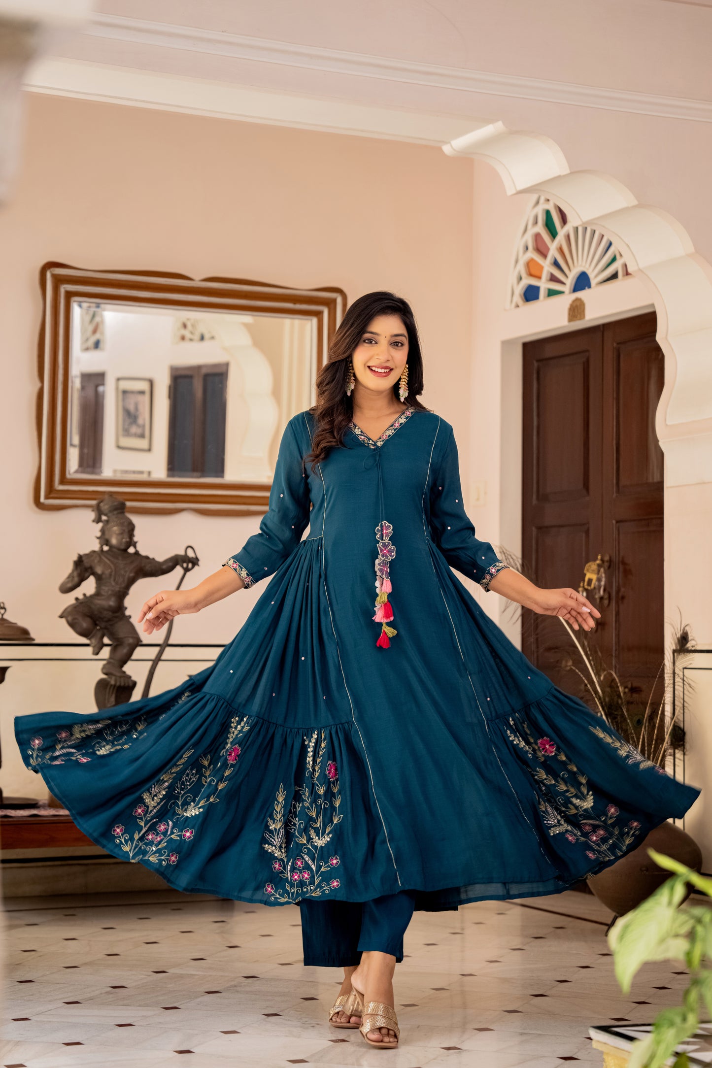 Woman in a blue embroidered traditional outfit standing in a decorated room.