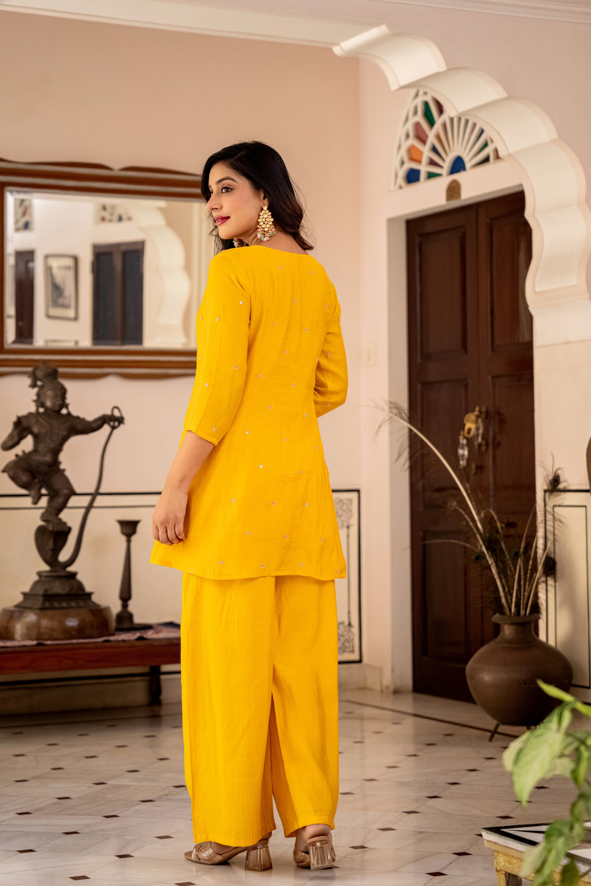 Woman in a yellow outfit standing in a decorated room with statues and plants.
